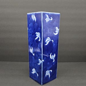 May include: A square, cobalt blue ceramic vase with a textured surface and white floral designs. The vase is a rectangular prism shape, with a dark blue background and white floral accents. The vase is approximately 8 inches tall.