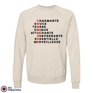 May include: A beige sweatshirt with black text that spells out "CONNASSE" using the first letter of each word in the list: "charmante", "douce", "tendre", "unique", "attachante", "surprenante", "essentielle", "merveilleuse".
