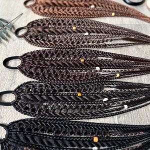 May include: Five hair extensions attached to black elastic hair ties, arranged in a gradient from light brown to black. Each extension features intricate braided strands, twisted sections, and small decorative beads in yellow and white colors.