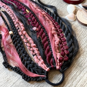 May include: A hair accessory featuring a collection of black, dusty pink, and deep burgundy synthetic hair strands, braids, and twists attached to a black elastic hair tie. The piece includes decorative elements like small gold beads and wooden beads.