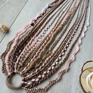 Könnte beinhalten: Geflochtene Haarverlängerungen in Braun-, Rosa- und Beigetönen, befestigt mit einem braunen Haargummi. Die Extensions weisen verschiedene Flechtmuster und Texturen auf, wobei einige Strähnen mit dekorativen Elementen verziert sind.