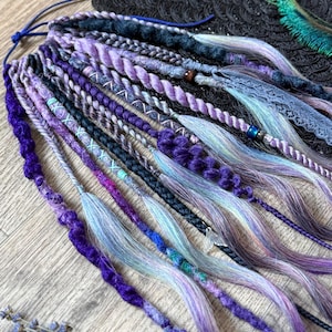 Könnte beinhalten: Handgefertigte Dreadlocks in Lila-, Blau- und Grautönen. Die Dreadlocks weisen verschiedene Texturen auf, darunter Zöpfe, Drehungen und glatte Abschnitte. Einige sind mit Perlen und Spitze verziert, was einen einzigartigen und künstlerischen Look erzeugt. Die Dreadlocks sind auf einer dunklen Oberfläche angeordnet.