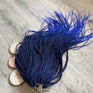 Könnte beinhalten: Ein Bündel dunkelblauer synthetischer Zopfhaare, eng zu einzelnen Strähnen geflochten. Das Haar ist mit einem natürlichen Bindfaden zusammengebunden. Die Zöpfe sind auf einer hellgrauen Holzoberfläche angeordnet.