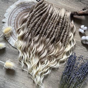May include: A bundle of wavy, light brown and blonde dreadlocks. The dreadlocks are arranged on a decorative, round, fringed placemat. Dried lavender, cotton buds, and dried thistle flowers are also present.