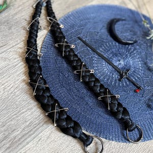 May include: Two long, thick black hair braids adorned with silver-toned metal safety pins arranged in a crisscross pattern. Each braid terminates in a circular metal ring at the bottom. A black sword-shaped hair accessory with a red gem is nearby.