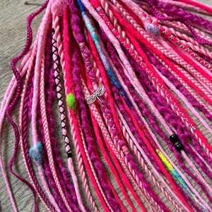 May include: A collection of vibrant, handmade dreadlocks in shades of pink and purple. The dreads feature various textures, including braids, twists, and wrapped sections. Embellishments include a silver dragonfly charm, beads, and colorful thread accents. The dreadlocks are tied together with a pink lace.