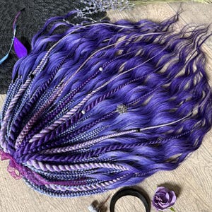 Puede incluir: Un conjunto de rastas moradas y lavanda, con secciones trenzadas y retorcidas. Adornado con cuentas plateadas y elementos decorativos. Una media luna negra, una flor morada y ramitas de lavanda están dispuestas cerca.