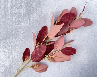 May include: A sprig of faux eucalyptus leaves in shades of pink and burgundy. The leaves are arranged in a cluster, with some leaves overlapping others. The leaves are on a light gray background.