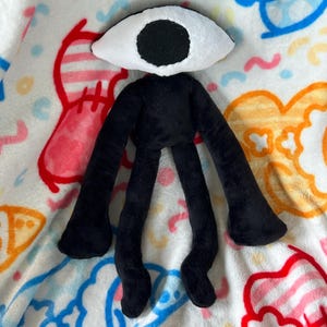 May include: A black plush toy with a large white eye and black pupil, long arms and legs. The toy is made of soft fabric and is set against a colourful patterned background.