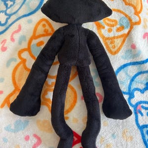 May include: A black plush doll with a rounded head, long arms, and long legs. The doll is made of a soft, fuzzy material. The doll's arms and legs are thin and elongated, and the feet are rounded. The doll is lying on a patterned blanket.