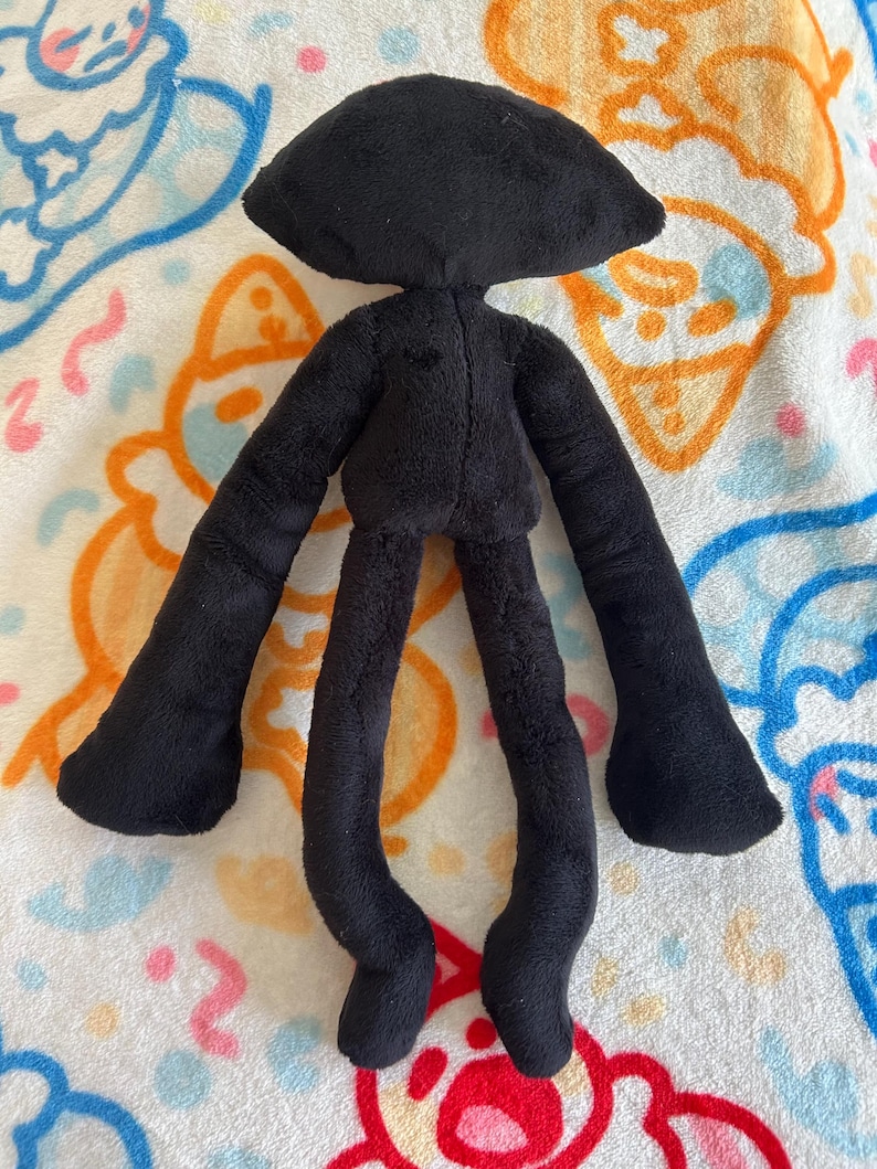 May include: A black plush doll with a rounded head, long arms, and long legs. The doll is made of a soft, fuzzy material. The doll's arms and legs are thin and elongated, and the feet are rounded. The doll is lying on a patterned blanket.