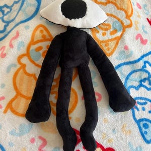 May include: A plush doll with a black body and long limbs. The doll has a large, white, eye-shaped head with a black pupil. The doll is lying on a patterned blanket with cartoon figures in blue, orange, and pink.