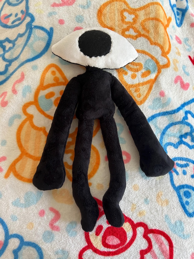 May include: A plush doll with a black body and long limbs. The doll has a large, white, eye-shaped head with a black pupil. The doll is lying on a patterned blanket with cartoon figures in blue, orange, and pink.