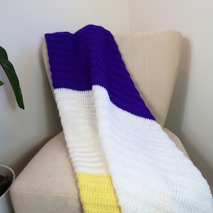 May include: A handmade crochet blanket draped over a beige chair. The blanket features a color block design with sections of purple, white, and yellow. The texture of the crochet stitches is visible.
