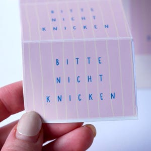 May include: A pink card with the text "BITTE NICHT KNICKEN" in blue. The card is held by a hand with a light pink nail polish. The card has vertical stripes and is folded.