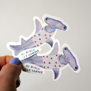 May include: Two hammerhead shark stickers. The sharks are grey with white bellies and red heart patterns. Each sticker has the German text "DU BISCH DER HAMMR" on a white banner.