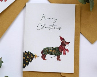 May include: Christmas greeting card featuring a dachshund wearing a festive sweater and Santa hat, pulling a Christmas tree. The card reads "Merry Christmas" in elegant script. Includes a brown envelope and sprigs of greenery.