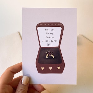 Könnte beinhalten: Eine Grußkarte mit einem Kaffee-Themen-Design. Die Illustration zeigt eine Ringbox mit einem Kaffeebohnenring und dem Text "Willst du mein ewiges Kaffeedate sein? (bitte)". Die Karte wird von einer Person gehalten.