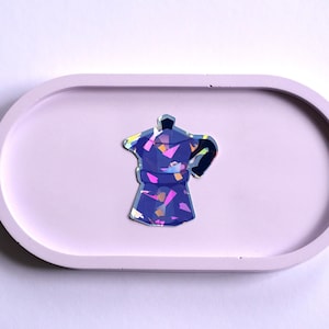 Könnte beinhalten: Ein dekoratives, abstraktes Kaffeekannen-Design in Blau-, Rosa- und Gelbtönen liegt auf einem hellvioletten, ovalen Tablett. Das Tablett besteht aus einem festen Material und befindet sich vor einem weißen Hintergrund. Die Kaffeekanne hat ein glänzendes, irisierendes Finish.