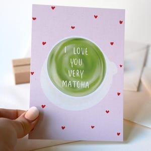 Könnte beinhalten: Eine hellrosa Grußkarte mit kleinen roten Herzen. Die Karte zeigt ein grünes Matcha-Tee-Design und die weißen Worte „I LOVE YOU VERY MATCHA“. Die Karte wird von einer Person gehalten.