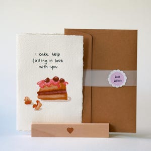 Könnte beinhalten: Eine Grußkarte mit dem Text "I cake help falling in love with you" und einer Kuchenillustration. Die Karte wird mit einem Holzständer und einem braunen Umschlag präsentiert.