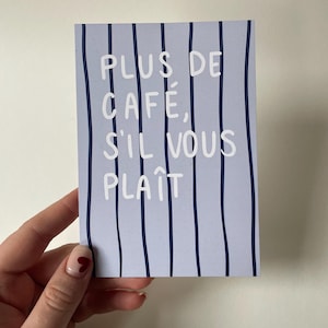 Könnte beinhalten: Eine hellblaue Karte mit vertikalen marineblauen Streifen. Die Karte zeigt den weißen Text "PLUS DE CAFE, S'IL VOUS PLAIT". Die Karte wird von einer Hand mit einem herzförmigen Nageldesign gehalten.