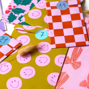 May include: A collection of colorful paper envelopes with various patterns, including smiley faces, a checkerboard, and holly berries. Each envelope has a numbered sticker. The color palette includes pink, green, orange, and gold.