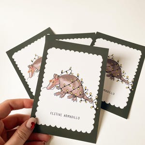 Könnte beinhalten: Vier Grußkarten mit einer Illustration eines festlichen Gürteltiers. Das Gürteltier ist mit Lichterketten und einem floralen Kopfschmuck geschmückt. Die Karten haben einen dunkelgrünen Rand mit einem gewellten Rand und dem Text "FESTIVE ARMADILLO".