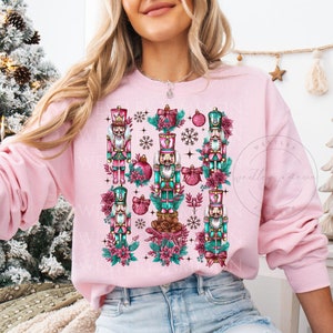 May include: Pink sweatshirt with a festive design featuring multiple nutcracker figures, ornaments, and floral elements. The design is centered on the front of the sweatshirt.