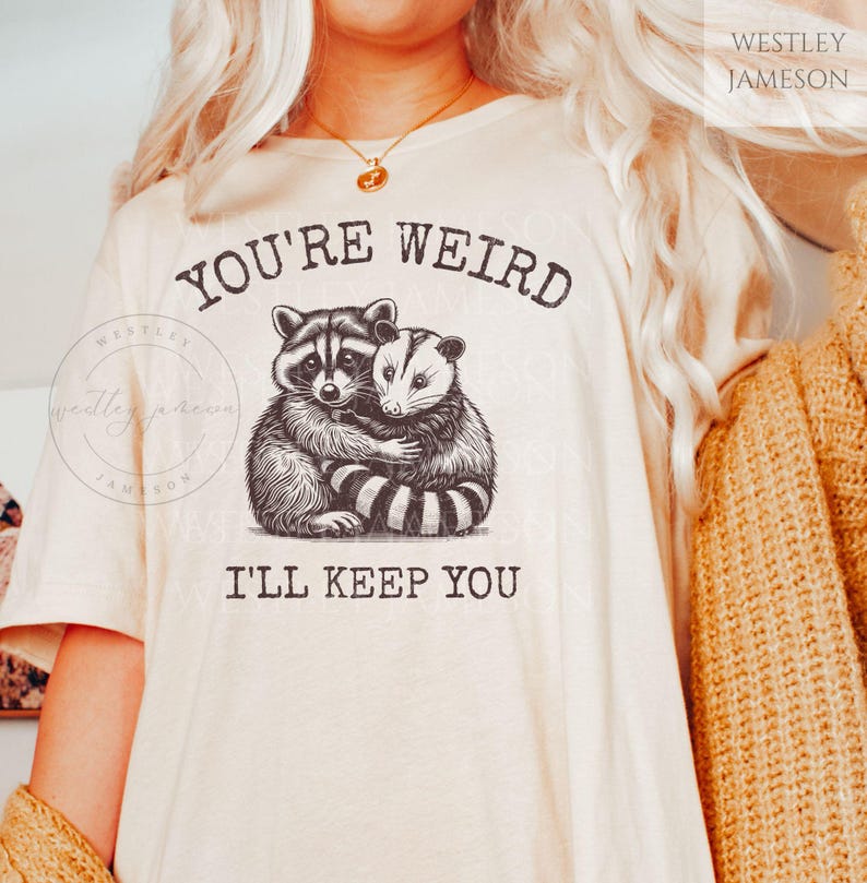 K&ouml;nnte beinhalten: Ein wei&szlig;es T-Shirt mit dem Aufdruck "You're Weird I'll Keep You". Der Text ist in schwarzer Schrift. Es gibt eine Schwarzwei&szlig;illustration eines Waschb&auml;ren und eines Opossums, die sich umarmen.