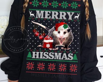 May include: Black Christmas sweater with a possum wearing a Santa hat and holding Christmas ornaments. The sweater has the text "Merry Hisssmas" and a green and red snowflake pattern.