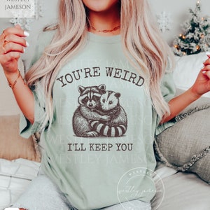 K&ouml;nnte beinhalten: Ein hellgr&uuml;nes T-Shirt mit einem Aufdruck eines Waschb&auml;ren und eines Opossums, die sich umarmen. Der Text "You're Weird I'll Keep You" ist auf das T-Shirt gedruckt.