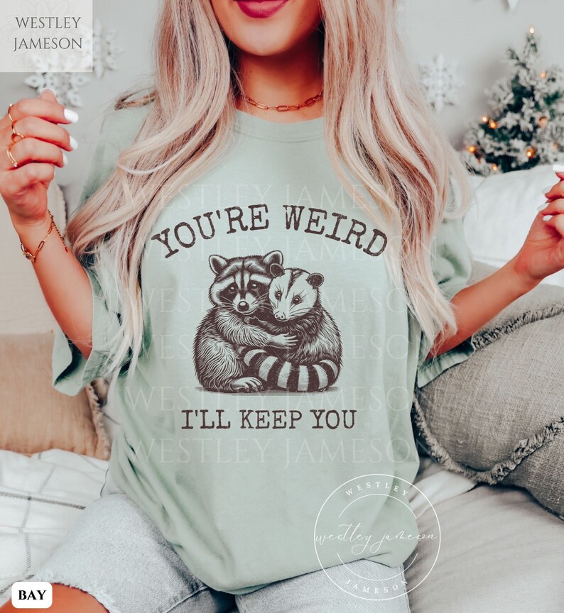 K&ouml;nnte beinhalten: Ein hellgr&uuml;nes T-Shirt mit einem Aufdruck eines Waschb&auml;ren und eines Opossums, die sich umarmen. Der Text "You're Weird I'll Keep You" ist auf das T-Shirt gedruckt.