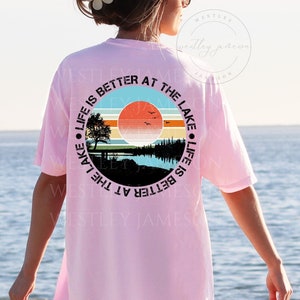 May include: A pink t-shirt with a black circle graphic on the back. The graphic features a sunset over a lake with trees and a bench. The text around the graphic reads "Life is better at the lake. Life is better at the lake."