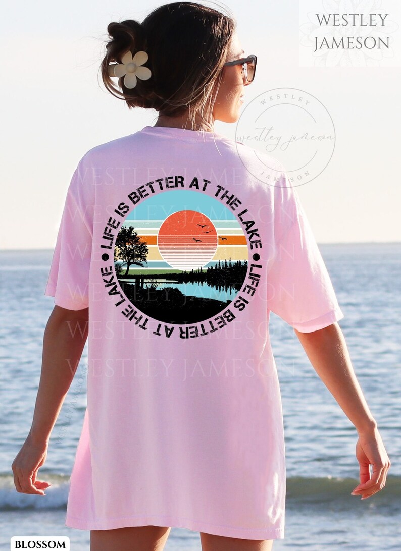 May include: A pink t-shirt with a black circle graphic on the back. The graphic features a sunset over a lake with trees and a bench. The text around the graphic reads "Life is better at the lake. Life is better at the lake."