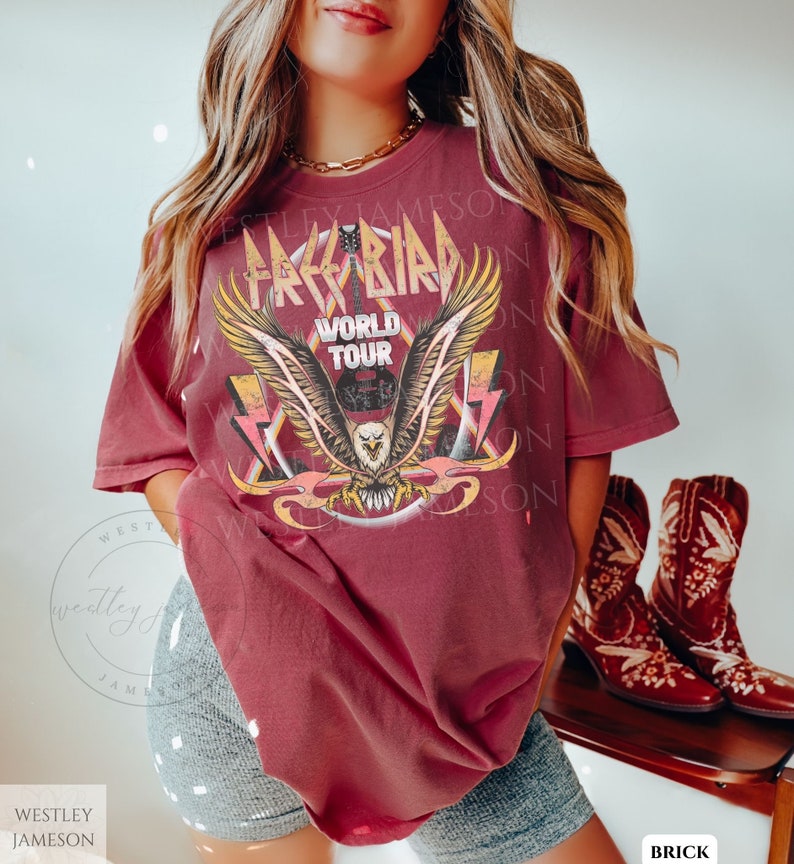 Pu&ograve; includere: Una maglietta color granata con una grafica di un'aquila con le ali spiegate, che tiene una chitarra. La grafica dice "Freebird World Tour".