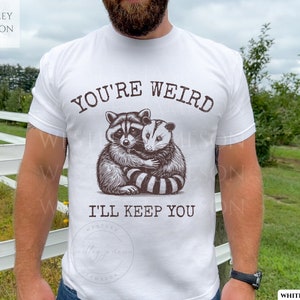K&ouml;nnte beinhalten: Wei&szlig;es T-Shirt mit einem Aufdruck eines Waschb&auml;ren und eines Opossums, die sich umarmen. Der Text "You're Weird I'll Keep You" ist auf das T-Shirt gedruckt.