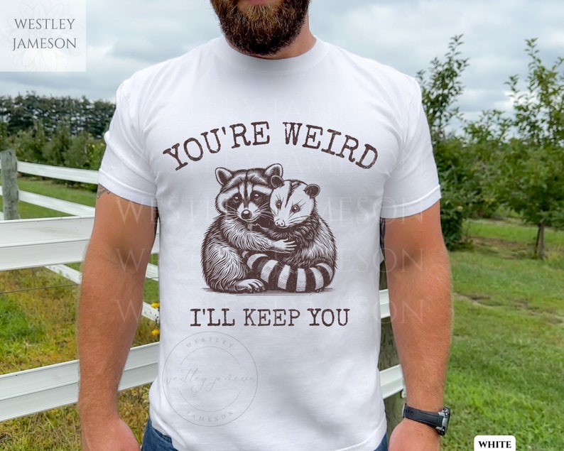 K&ouml;nnte beinhalten: Wei&szlig;es T-Shirt mit einem Aufdruck eines Waschb&auml;ren und eines Opossums, die sich umarmen. Der Text "You're Weird I'll Keep You" ist auf das T-Shirt gedruckt.