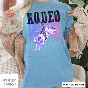 Puede incluir: Una camiseta azul claro con la palabra "RODEO" impresa en letras negras. El dise&ntilde;o presenta una silueta morada de un vaquero montando un toro sobre un fondo morado y azul. El texto "WESTLEYJAMESON" est&aacute; impreso en blanco en la parte inferior de la camiseta. La camiseta est&aacute; etiquetada como "WASHED DENIM".