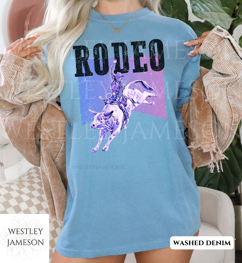 Puede incluir: Una camiseta azul claro con la palabra "RODEO" impresa en letras negras. El dise&ntilde;o presenta una silueta morada de un vaquero montando un toro sobre un fondo morado y azul. El texto "WESTLEYJAMESON" est&aacute; impreso en blanco en la parte inferior de la camiseta. La camiseta est&aacute; etiquetada como "WASHED DENIM".