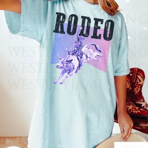 Puede incluir: Una camiseta azul claro con la palabra "RODEO" impresa en negro con un efecto desgastado. El gr&aacute;fico presenta una silueta morada de un vaquero montando un toro sobre un fondo rosa y morado.