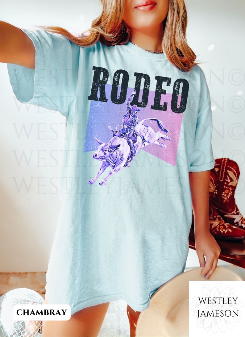Puede incluir: Una camiseta azul claro con la palabra "RODEO" impresa en negro con un efecto desgastado. El gr&aacute;fico presenta una silueta morada de un vaquero montando un toro sobre un fondo rosa y morado.