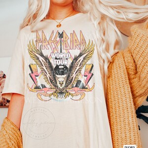 Pu&ograve; includere: Una maglietta color crema con un design grafico vintage che presenta un'aquila calva con le ali spiegate, una chitarra e il testo "Free Bird World Tour". La grafica &egrave; in tonalit&agrave; rosa, giallo e blu.