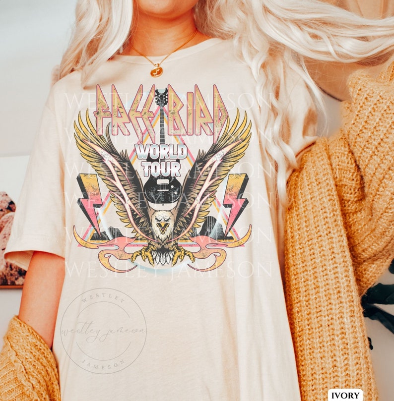 Pu&ograve; includere: Una maglietta color crema con un design grafico vintage che presenta un'aquila calva con le ali spiegate, una chitarra e il testo "Free Bird World Tour". La grafica &egrave; in tonalit&agrave; rosa, giallo e blu.