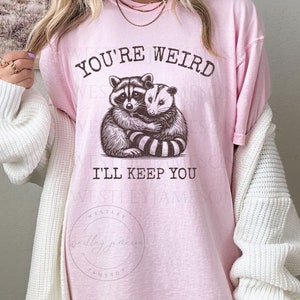 K&ouml;nnte beinhalten: Pinkes T-Shirt mit einem Aufdruck eines Waschb&auml;ren und einer Opossum, die sich umarmen. Der Text "You're Weird I'll Keep You" ist auf dem T-Shirt aufgedruckt.