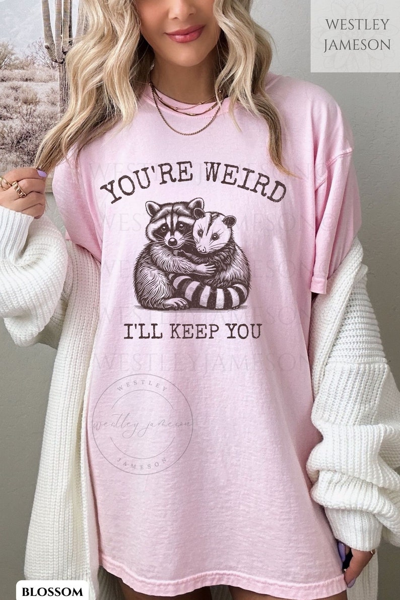 K&ouml;nnte beinhalten: Pinkes T-Shirt mit einem Aufdruck eines Waschb&auml;ren und einer Opossum, die sich umarmen. Der Text "You're Weird I'll Keep You" ist auf dem T-Shirt aufgedruckt.