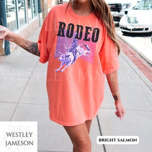 Puede incluir: Una camiseta de color salm&oacute;n brillante con un gr&aacute;fico de un vaquero montando un toro. El gr&aacute;fico es de color morado y azul con un fondo rosa y morado. El texto "RODEO" est&aacute; impreso en negro en la camiseta.