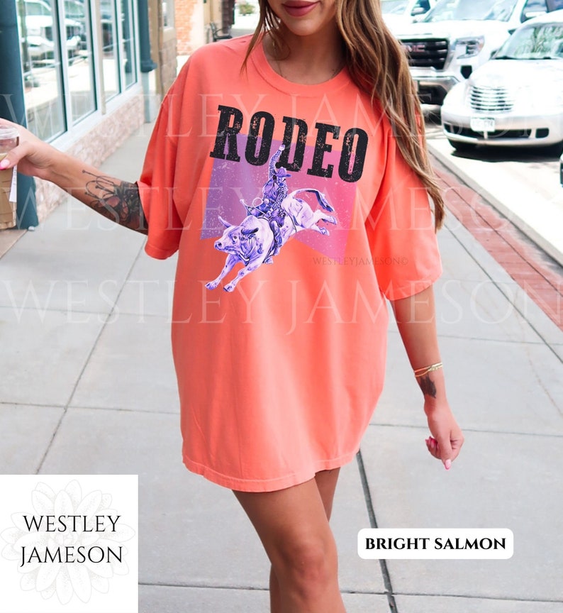 Puede incluir: Una camiseta de color salm&oacute;n brillante con un gr&aacute;fico de un vaquero montando un toro. El gr&aacute;fico es de color morado y azul con un fondo rosa y morado. El texto "RODEO" est&aacute; impreso en negro en la camiseta.