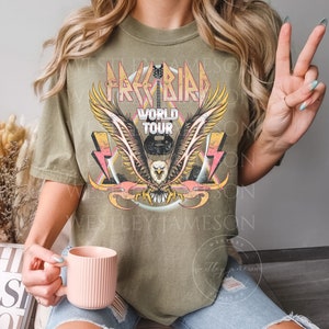 Pu&ograve; includere: Una maglietta verde kaki con una grafica vintage di un'aquila con le ali spiegate, una chitarra e il testo "Free Bird World Tour".