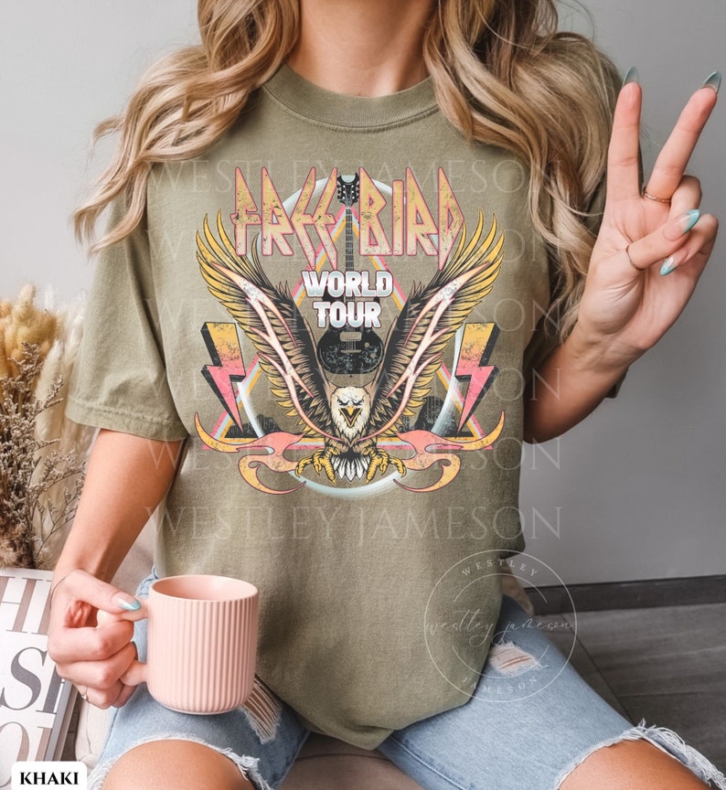 Pu&ograve; includere: Una maglietta verde kaki con una grafica vintage di un'aquila con le ali spiegate, una chitarra e il testo "Free Bird World Tour".
